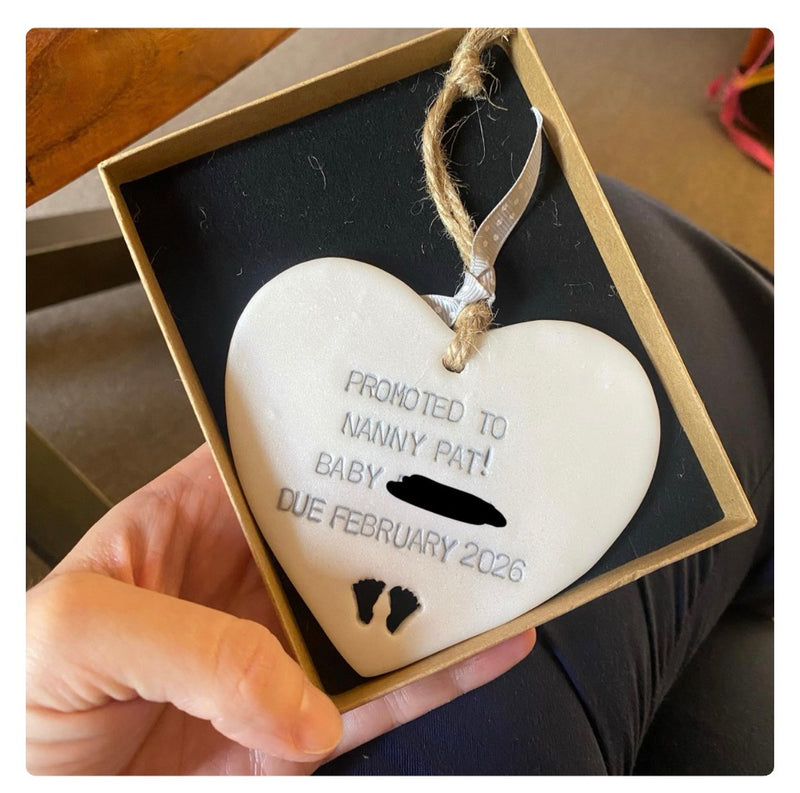 A hand holds an open gift box containing a Pearlised white polymer clay pregnancy reveal heart with an hand stamped message and small cut out footprints. The ornament hangs from a jute ribbon inside a tan box with a black insert. 