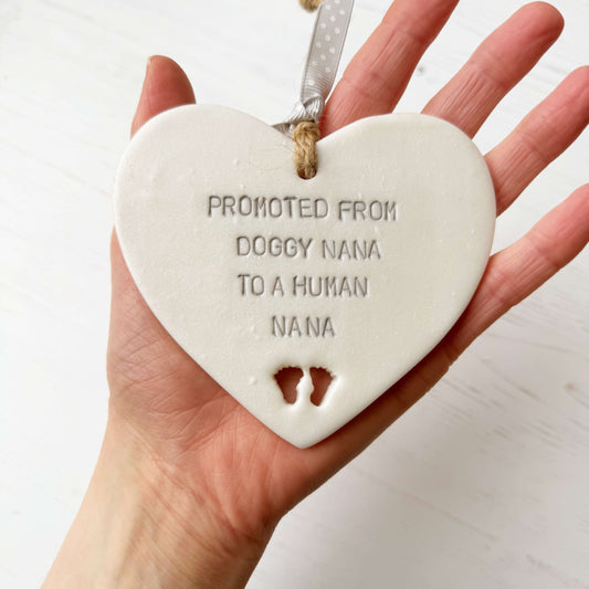 Pearlised white clay hanging heart with baby feet cut out of the bottom and grey personalisation, the heart is personalised with PROMOTED FROM DOGGY NANA TO A HUMAN NANA