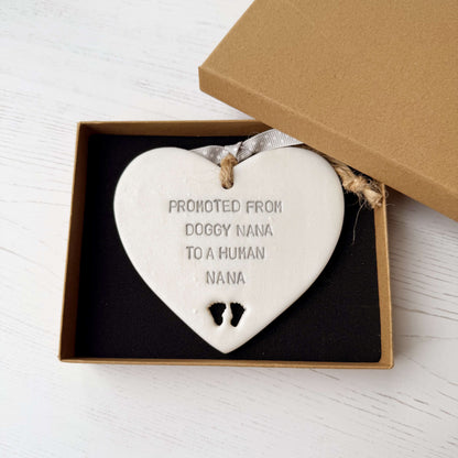 Pearlised white clay hanging heart with baby feet cut out of the bottom and grey personalisation, the heart is personalised with PROMOTED FROM DOGGY NANA TO A HUMAN NANA