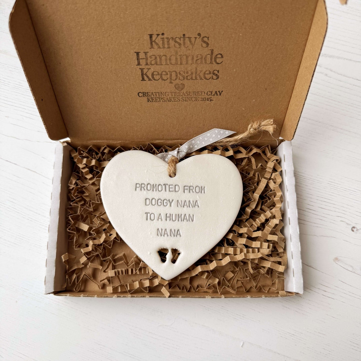 Pearlised white clay hanging heart with baby feet cut out of the bottom and grey personalisation, the heart is personalised with PROMOTED FROM DOGGY NANA TO A HUMAN NANA