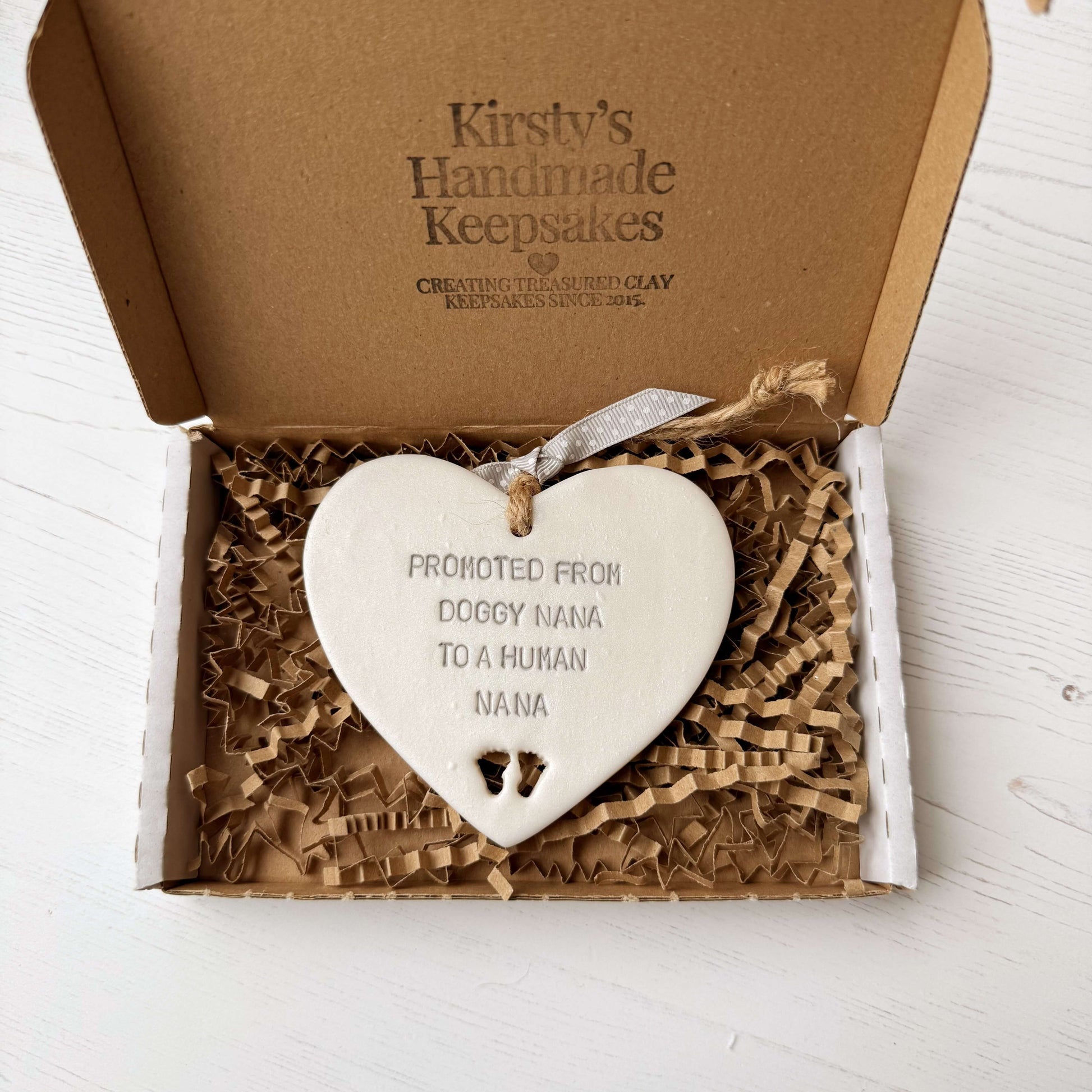 Pearlised white clay hanging heart with baby feet cut out of the bottom and grey personalisation, the heart is personalised with PROMOTED FROM DOGGY NANA TO A HUMAN NANA