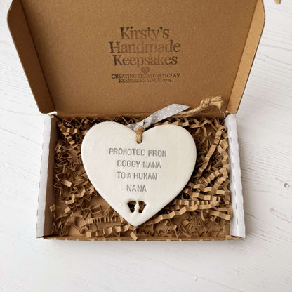 Pearlised white clay hanging heart with baby feet cut out of the bottom and grey personalisation, the heart is personalised with PROMOTED FROM DOGGY NANA TO A HUMAN NANA