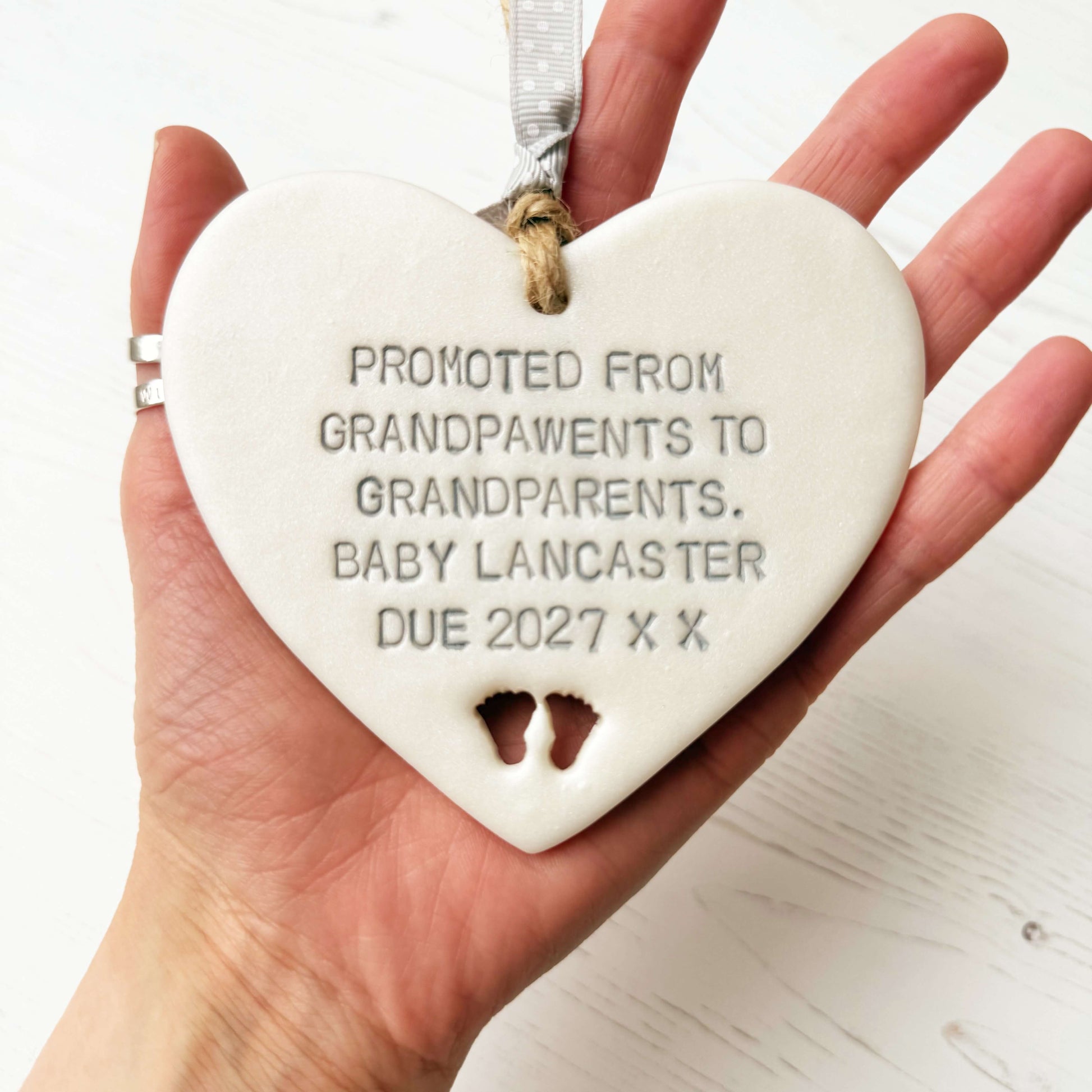Pearlised white clay hanging heart with baby feet cut out of the bottom and grey personalisation, the heart is personalised with PROMOTED FROM GRANDPAWENTS TO GRANDPARENTS. BABY LANCASTER DUE 2027 X X