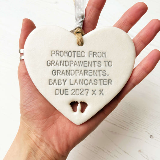 Pearlised white clay hanging heart with baby feet cut out of the bottom and grey personalisation, the heart is personalised with PROMOTED FROM GRANDPAWENTS TO GRANDPARENTS. BABY LANCASTER DUE 2027 X X