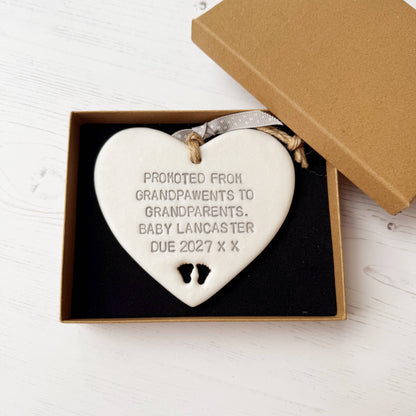 Pearlised white clay hanging heart with baby feet cut out of the bottom and grey personalisation, the heart is personalised with PROMOTED FROM GRANDPAWENTS TO GRANDPARENTS. BABY LANCASTER DUE 2027 X X