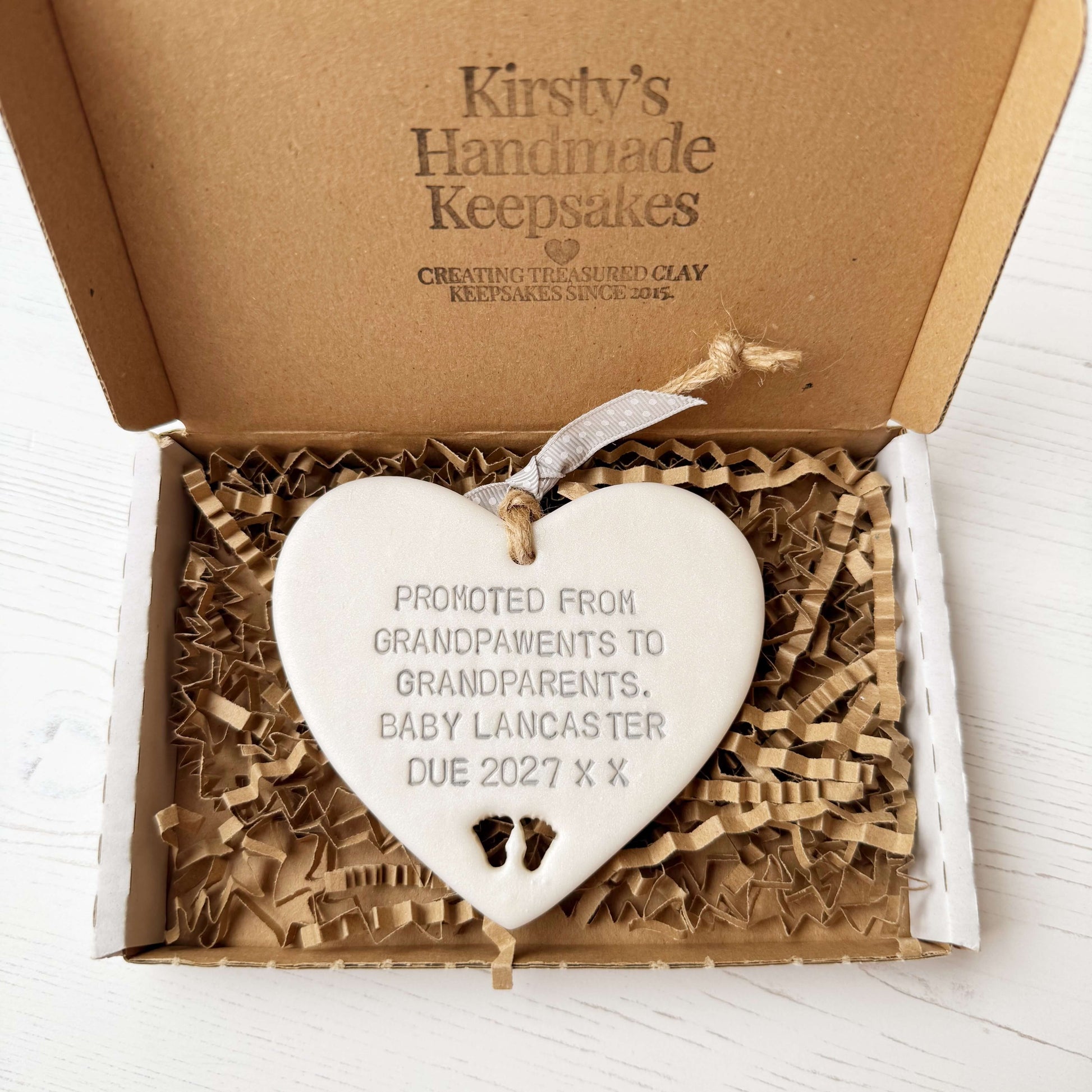 Pearlised white clay hanging heart with baby feet cut out of the bottom and grey personalisation, the heart is personalised with PROMOTED FROM GRANDPAWENTS TO GRANDPARENTS. BABY LANCASTER DUE 2027 X X