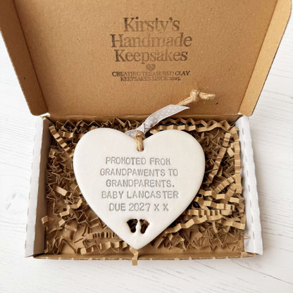 Pearlised white clay hanging heart with baby feet cut out of the bottom and grey personalisation, the heart is personalised with PROMOTED FROM GRANDPAWENTS TO GRANDPARENTS. BABY LANCASTER DUE 2027 X X