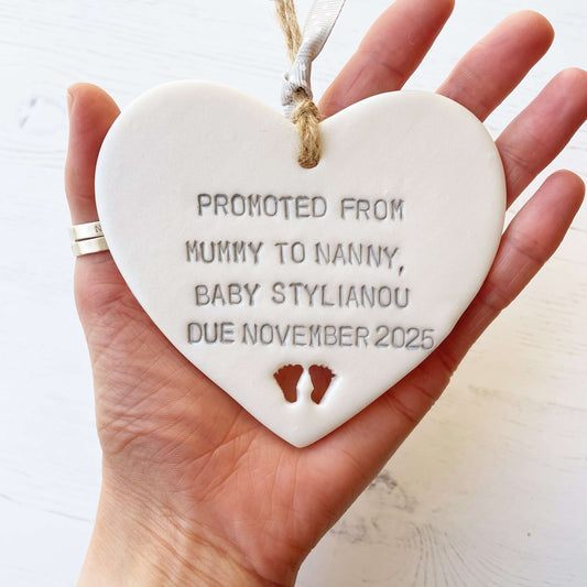 Pearlised white clay hanging heart with baby feet cut out of the bottom and grey personalisation, the heart is personalised with PROMOTED FROM MUMMY TO NANNY, BABY STYLIANOU DUE NOVEMBER 2025