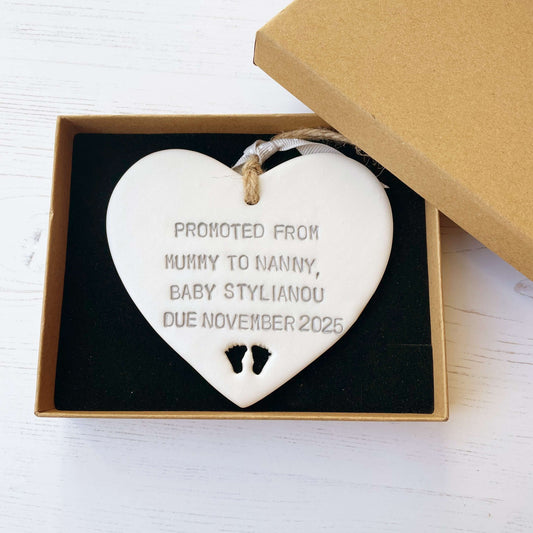 Pearlised white clay hanging heart with baby feet cut out of the bottom and grey personalisation, the heart is personalised with PROMOTED FROM MUMMY TO NANNY, BABY STYLIANOU DUE NOVEMBER 2025