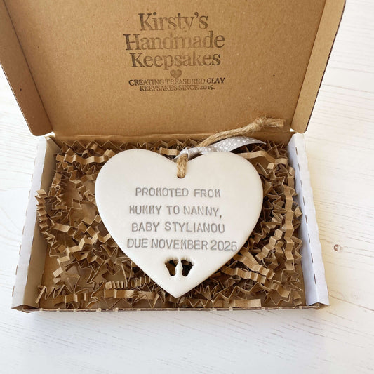 Pearlised white clay hanging heart with baby feet cut out of the bottom and grey personalisation, the heart is personalised with PROMOTED FROM MUMMY TO NANNY, BABY STYLIANOU DUE NOVEMBER 2025