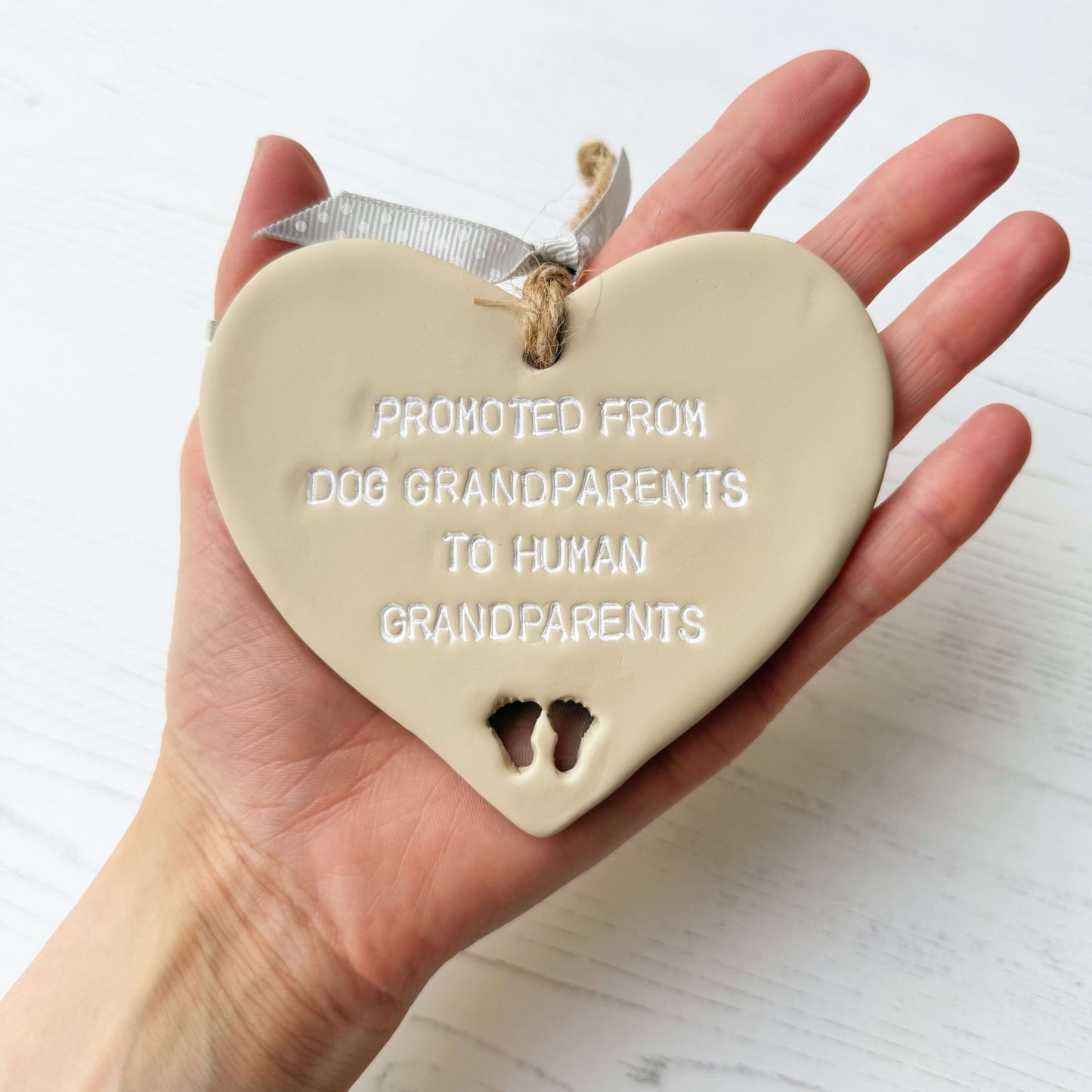 Beige clay hanging heart with baby feet cut out of the bottom and white personalisation, the heart is personalised with PROMOTED FROM DOG GRANDPARENTS TO HIMAN GRANDPARENTS