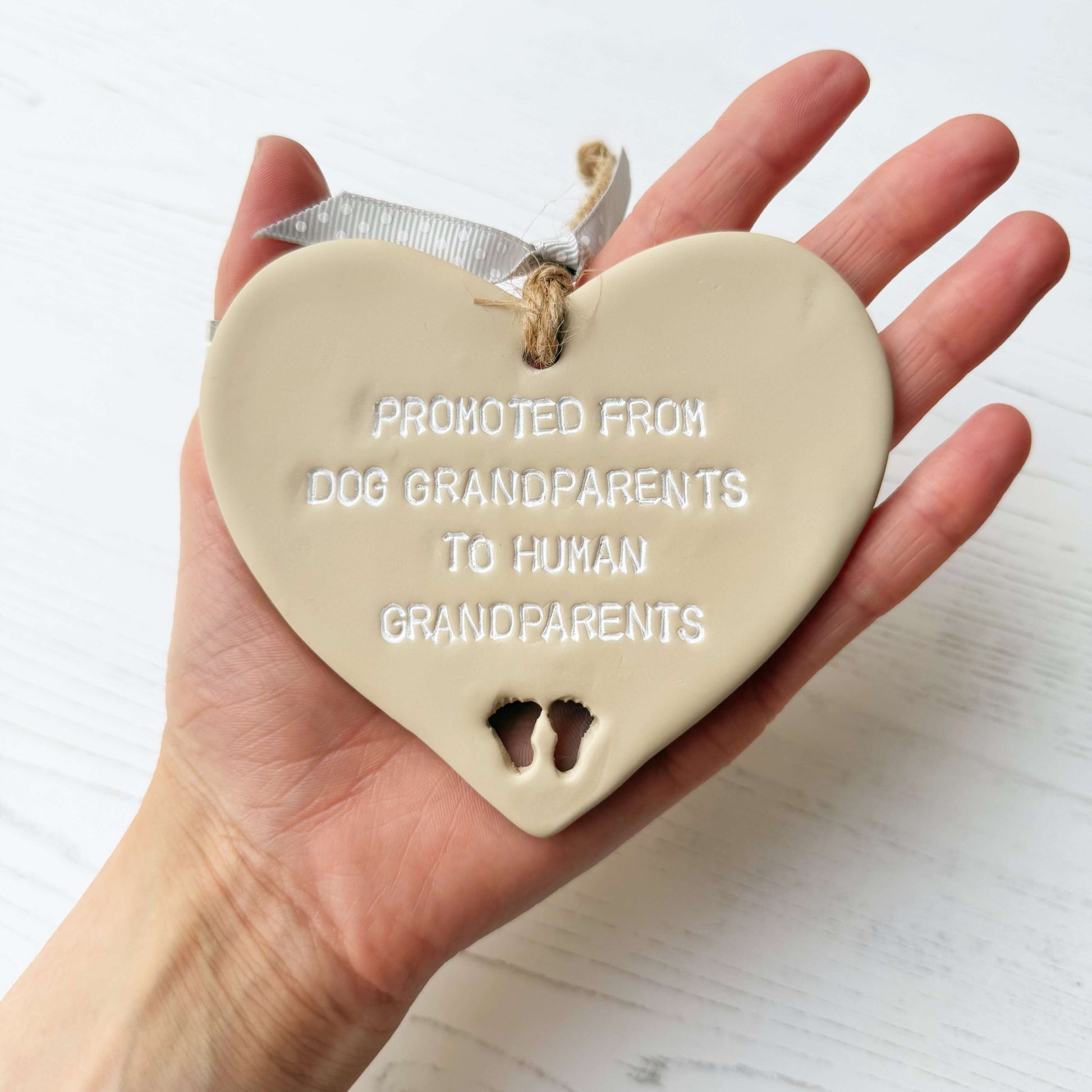 Beige clay hanging heart with baby feet cut out of the bottom and white personalisation, the heart is personalised with PROMOTED FROM DOG GRANDPARENTS TO HIMAN GRANDPARENTS