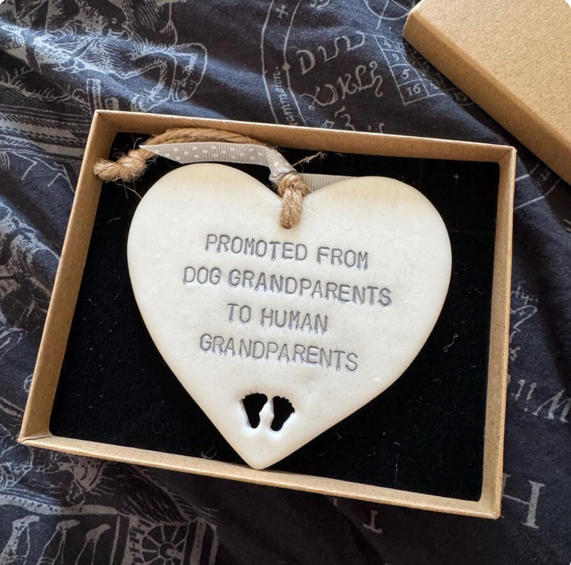 A Pearlised white polymer clay heart with PROMOTED FROM DOG GRANDPARENTS TO HUMAN GRANDPARENTS hand stamped on it, displayed inside its open gift box. The ornament is off-white with dark gray lettering and features two small footprints at the bottom, suspended by a twine loop.