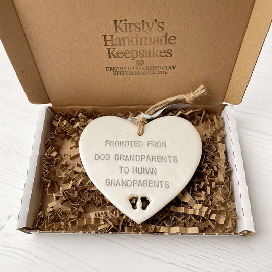 Pearlised white clay hanging heart with baby feet cut out of the bottom and grey personalisation, the heart is personalised with PROMOTED FROM DOG GRANDPARENTS TO HUMAN GRANDPARENTS
