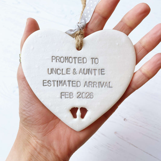 Pearlised white clay hanging heart with baby feet cut out of the bottom and grey personalisation, the heart is personalised with PROMOTED TO UNCLE & AUNTIE ESTIMATED ARRIVAL FEB 2026