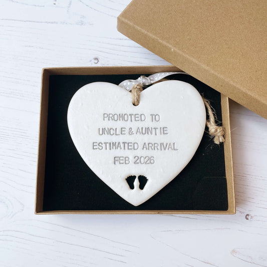 Pearlised white clay hanging heart with baby feet cut out of the bottom and grey personalisation, the heart is personalised with PROMOTED TO UNCLE & AUNTIE ESTIMATED ARRIVAL FEB 2026