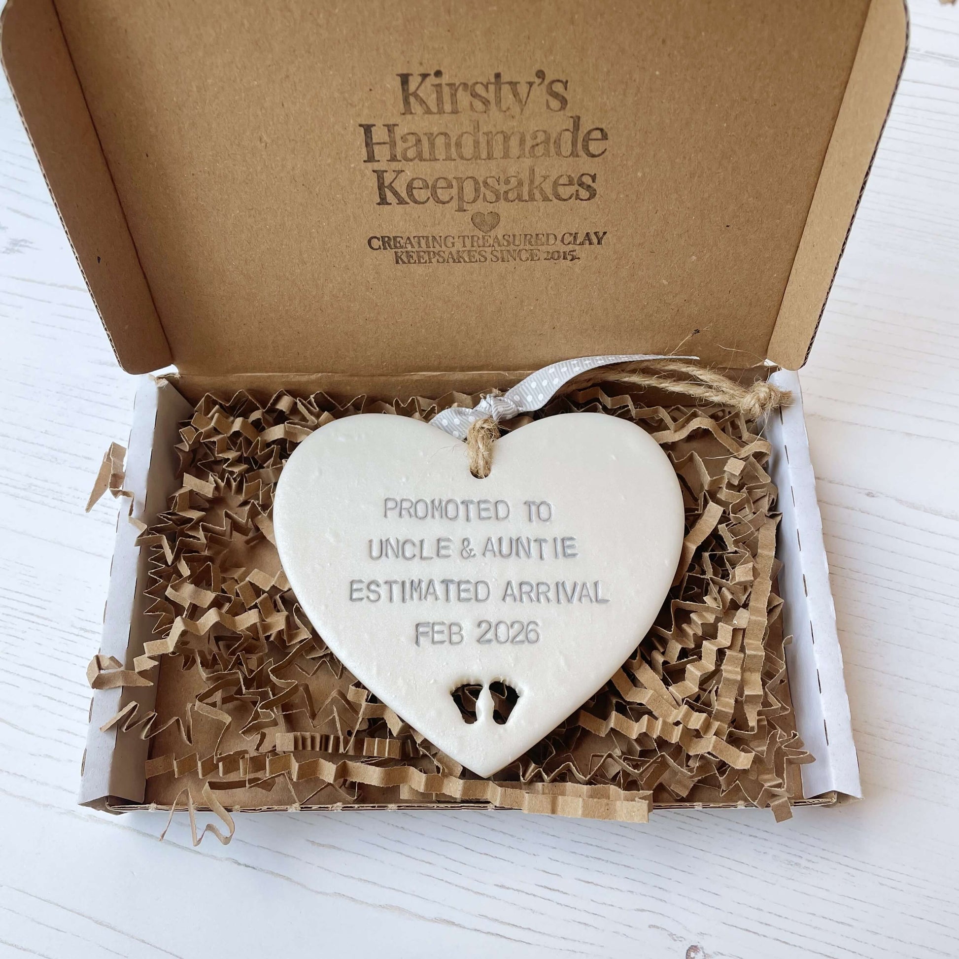Pearlised white clay hanging heart with baby feet cut out of the bottom and grey personalisation, the heart is personalised with PROMOTED TO UNCLE & AUNTIE ESTIMATED ARRIVAL FEB 2026