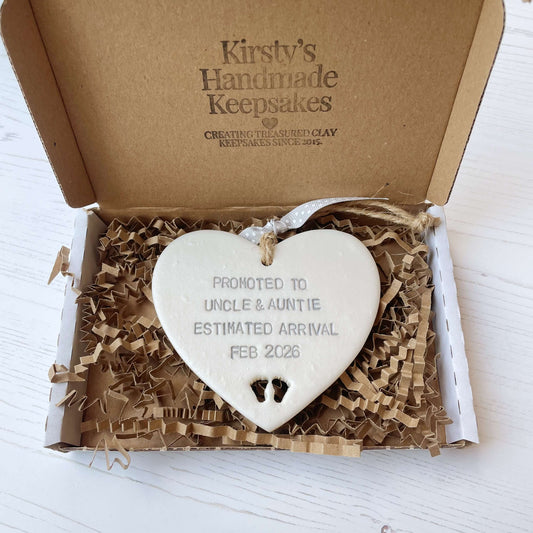 Pearlised white clay hanging heart with baby feet cut out of the bottom and grey personalisation, the heart is personalised with PROMOTED TO UNCLE & AUNTIE ESTIMATED ARRIVAL FEB 2026