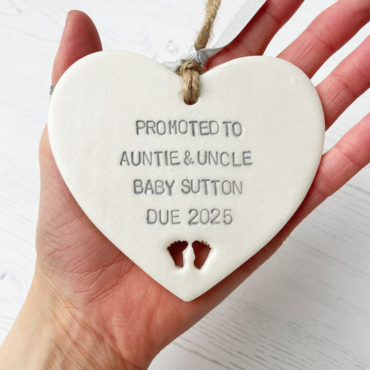 Pearlised white clay hanging heart with baby feet cut out of the bottom and grey personalisation, the heart is personalised with PROMOTED TO GREAT AUNTIE & UNCLE BABY SUTTON DUE 2025