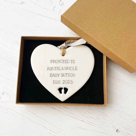 Pearlised white clay hanging heart with baby feet cut out of the bottom and grey personalisation, the heart is personalised with PROMOTED TO GREAT AUNTIE & UNCLE BABY SUTTON DUE 2025