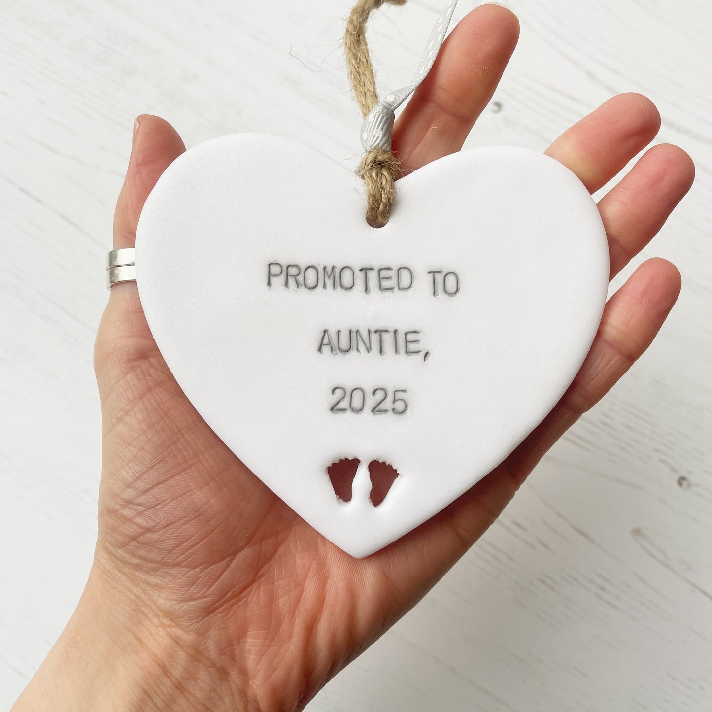 Pearlised white clay hanging heart with baby feet cut out of the bottom and grey personalisation, the heart is personalised with PROMOTED TO AUNTIE, 2025