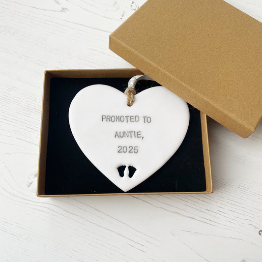 Pearlised white clay hanging heart with baby feet cut out of the bottom and grey personalisation, the heart is personalised with PROMOTED TO AUNTIE, 2025