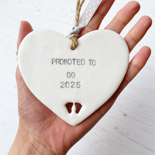 Pearlised white clay hanging heart with baby feet cut out of the bottom and grey personalisation, the heart is personalised with PROMOTED TO GG 2025