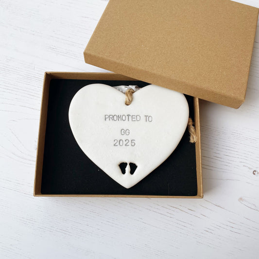 Pearlised white clay hanging heart with baby feet cut out of the bottom and grey personalisation, the heart is personalised with PROMOTED TO GG 2025