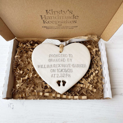 Pearlised white clay hanging heart with baby feet cut out of the bottom and grey personalisation, the heart is personalised with PROMOTED TO GRANDAD BY WILLIAM REX WAYE-BARKER ON 10/05/25 AT 2:32 PM