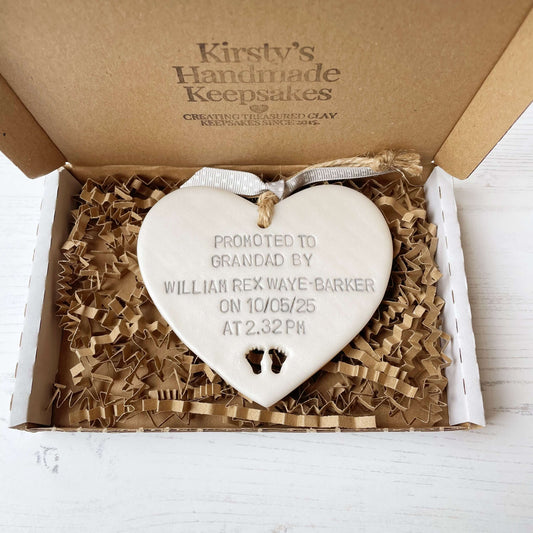 Pearlised white clay hanging heart with baby feet cut out of the bottom and grey personalisation, the heart is personalised with PROMOTED TO GRANDAD BY WILLIAM REX WAYE-BARKER ON 10/05/25 AT 2:32 PM