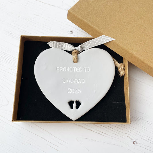 Grey clay hanging heart with baby feet cut out of the bottom and white personalisation, the heart is personalised with PROMOTED TO GRANDAD 2025