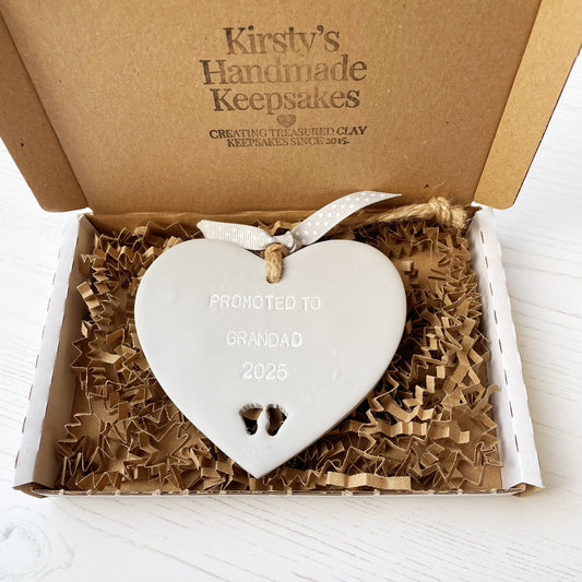 Grey clay hanging heart with baby feet cut out of the bottom and white personalisation, the heart is personalised with PROMOTED TO GRANDAD 2025