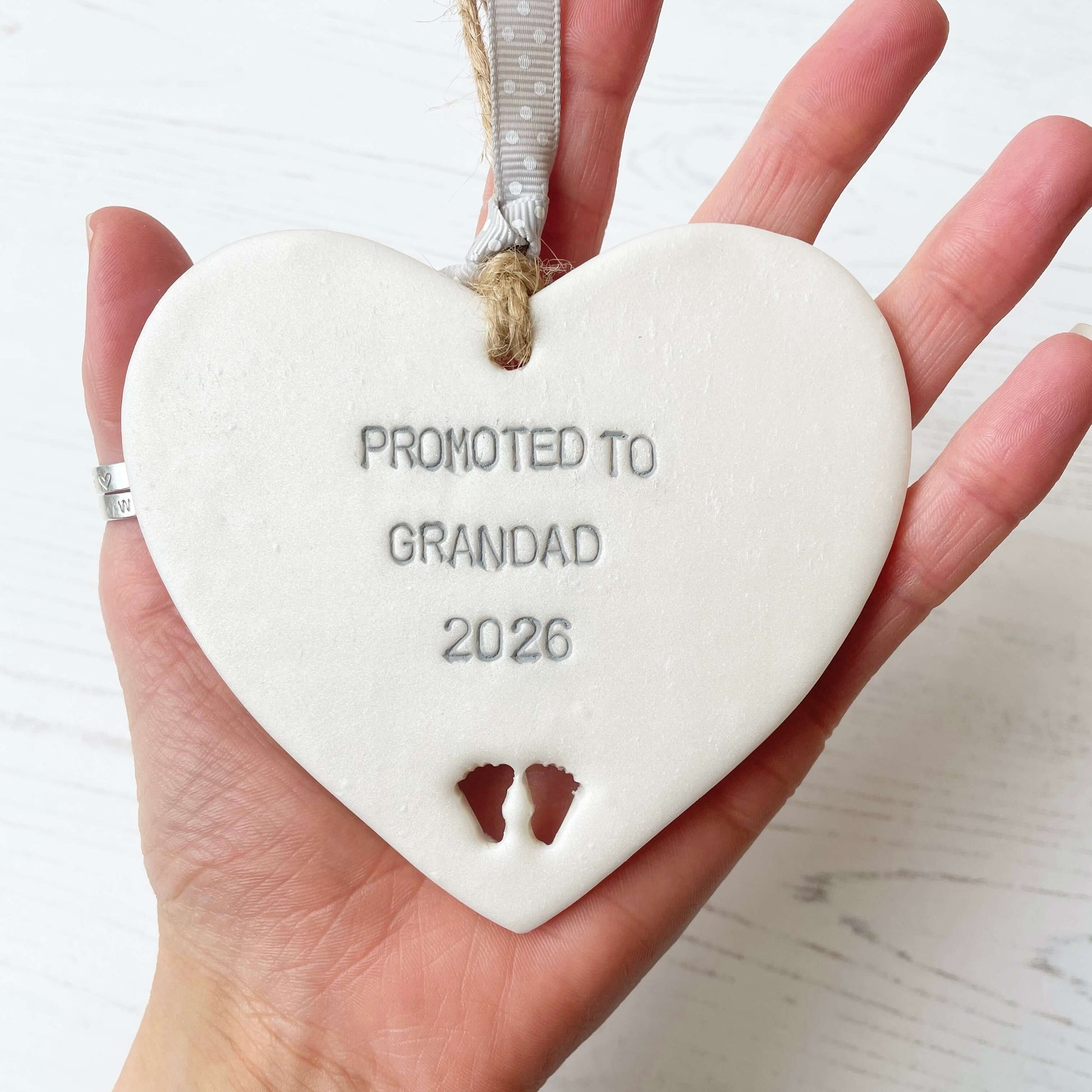 Pearlised white clay hanging heart with baby feet cut out of the bottom and grey personalisation, the heart is personalised with PROMOTED TO GRANDAD 2026