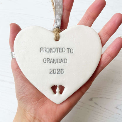 Pearlised white clay hanging heart with baby feet cut out of the bottom and grey personalisation, the heart is personalised with PROMOTED TO GRANDAD 2026