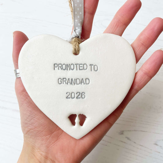 Pearlised white clay hanging heart with baby feet cut out of the bottom and grey personalisation, the heart is personalised with PROMOTED TO GRANDAD 2026