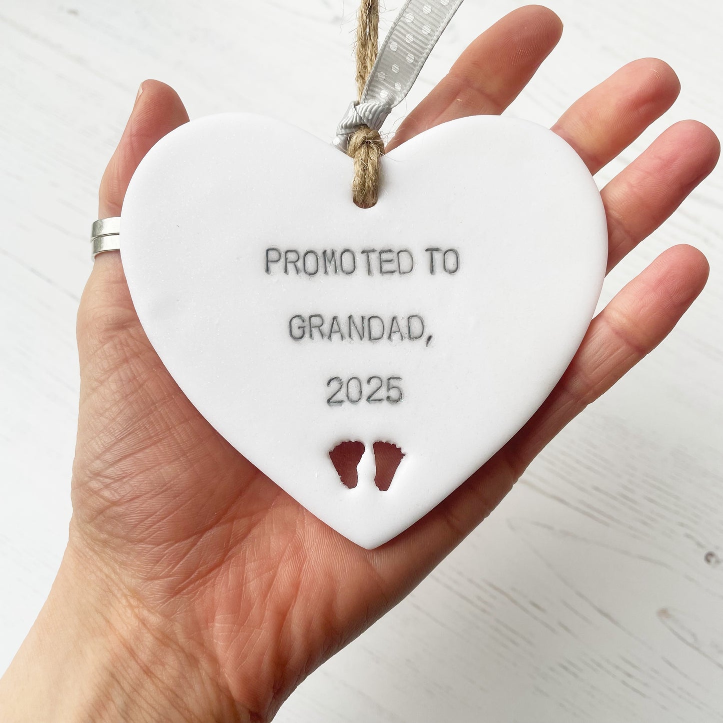 Pearlised white clay hanging heart with baby feet cut out of the bottom and grey personalisation, the heart is personalised with PROMOTED TO GRANDAD, 2025