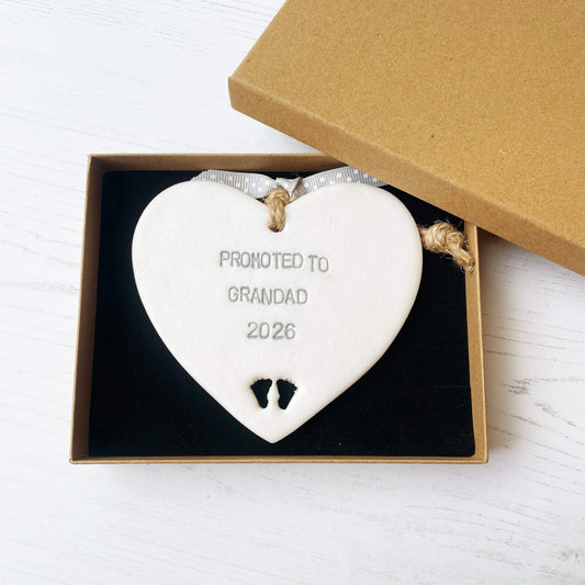 Pearlised white clay hanging heart with baby feet cut out of the bottom and grey personalisation, the heart is personalised with PROMOTED TO GRANDAD 2026