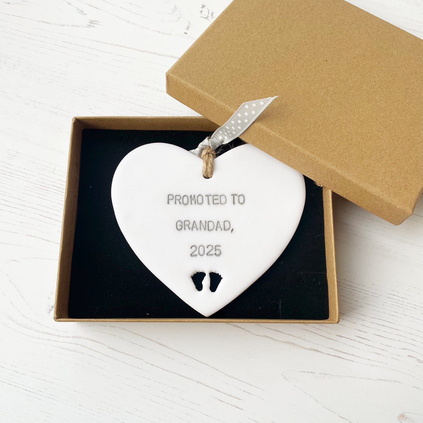 Pearlised white clay hanging heart with baby feet cut out of the bottom and grey personalisation, the heart is personalised with PROMOTED TO GRANDAD, 2025