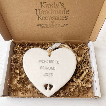 Pearlised white clay hanging heart with baby feet cut out of the bottom and grey personalisation, the heart is personalised with PROMOTED TO GRANDAD 2026
