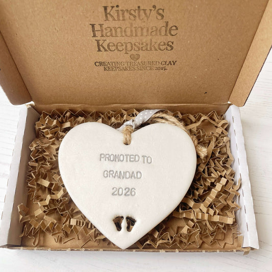Pearlised white clay hanging heart with baby feet cut out of the bottom and grey personalisation, the heart is personalised with PROMOTED TO GRANDAD 2026