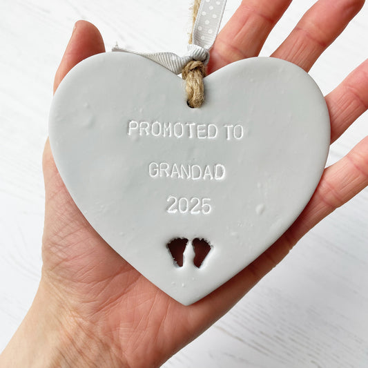 Grey clay hanging heart with baby feet cut out of the bottom and white personalisation, the heart is personalised with PROMOTED TO GRANDAD 2025