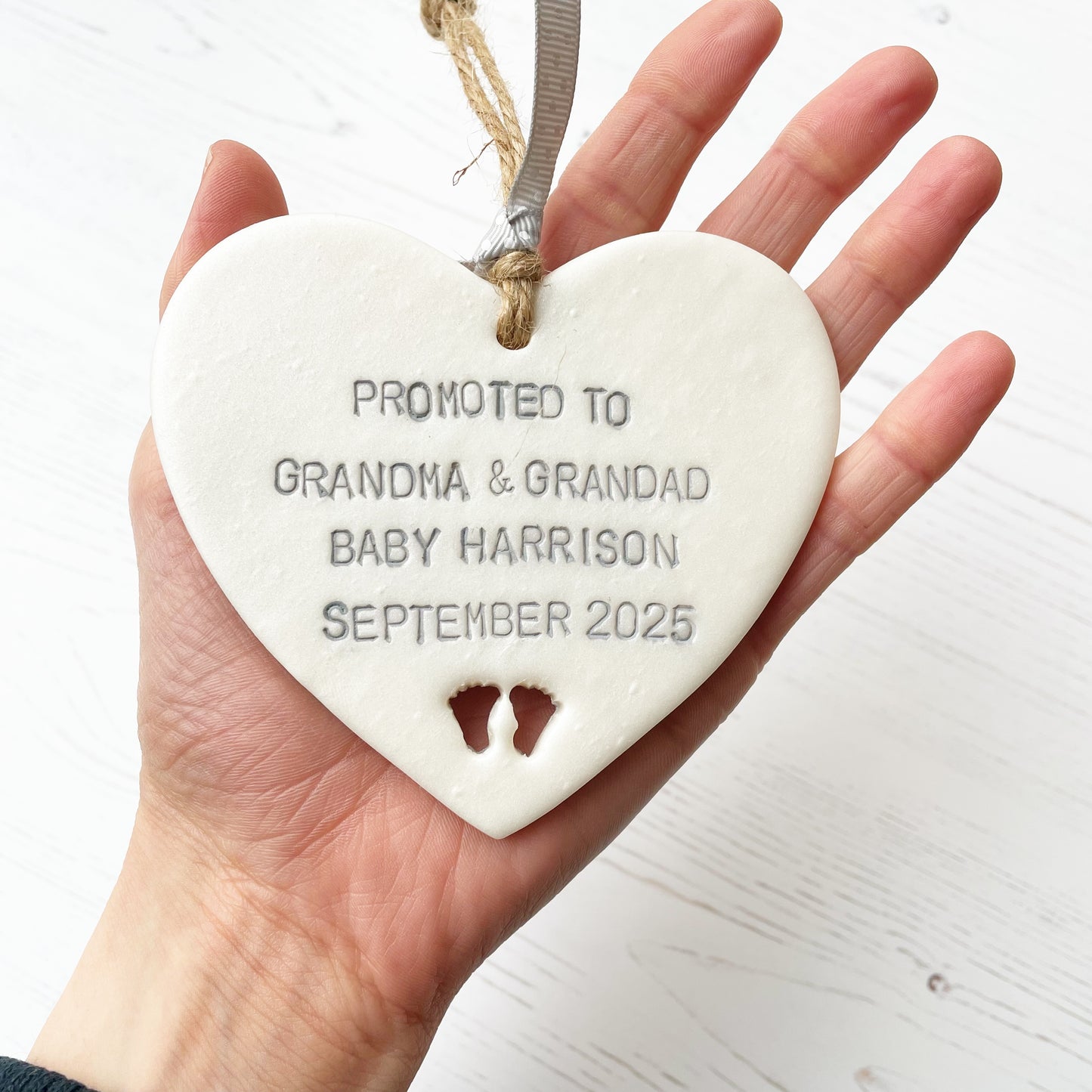 Pearlised white clay hanging heart with baby feet cut out of the bottom and grey personalisation, the heart is personalised with PROMOTED TO GRANDMA & GRANDAD BABY HARRISON SEPTEMBER 2025