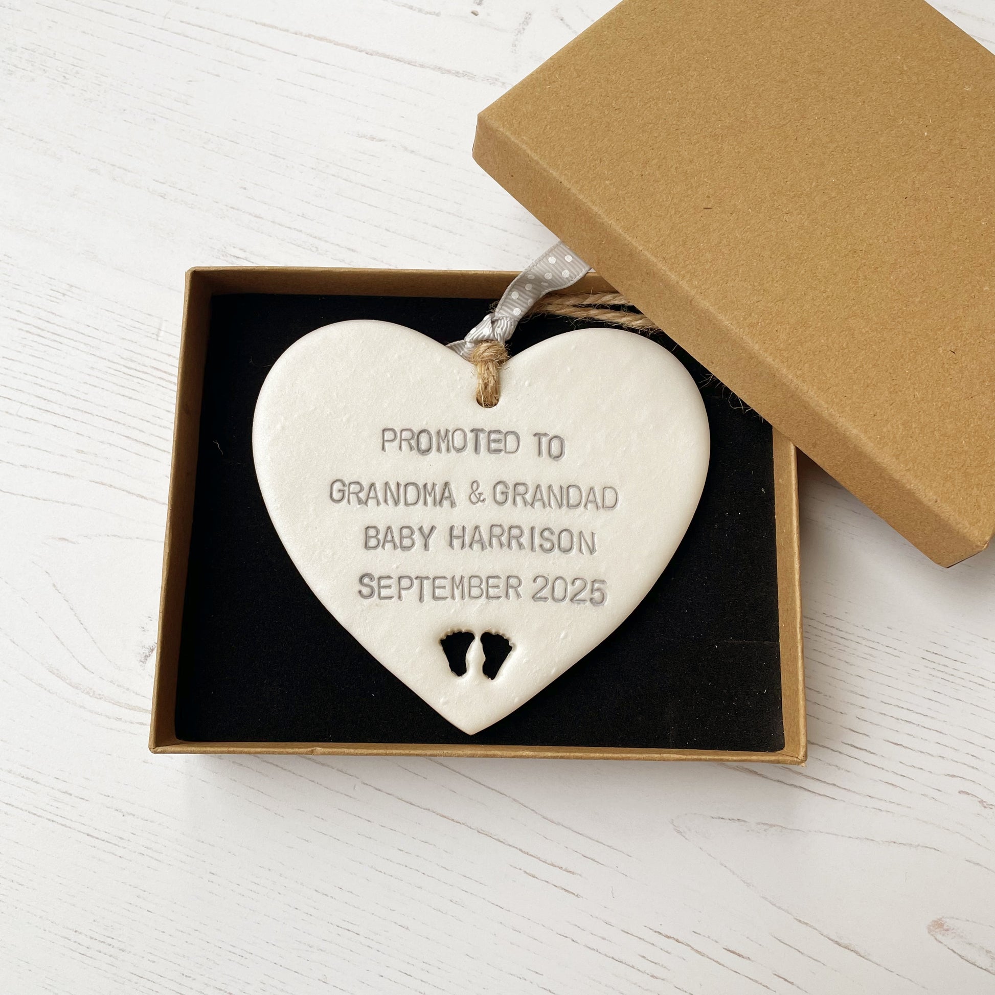 Pearlised white clay hanging heart with baby feet cut out of the bottom and grey personalisation, the heart is personalised with PROMOTED TO GRANDMA & GRANDAD BABY HARRISON SEPTEMBER 2025