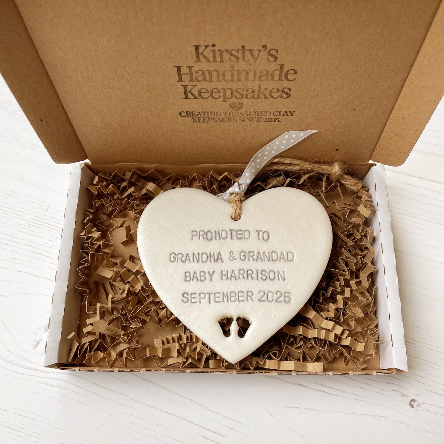 Pearlised white clay hanging heart with baby feet cut out of the bottom and grey personalisation, the heart is personalised with PROMOTED TO GRANDMA & GRANDAD BABY HARRISON SEPTEMBER 2025