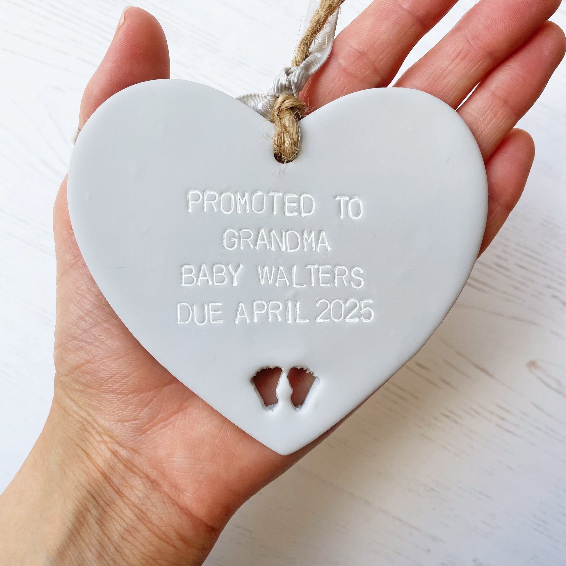 Grey clay hanging heart with baby feet cut out of the bottom and white personalisation, the heart is personalised with PROMOTED TO GRANDMA BABY WALTERS DUE APRIL 2025