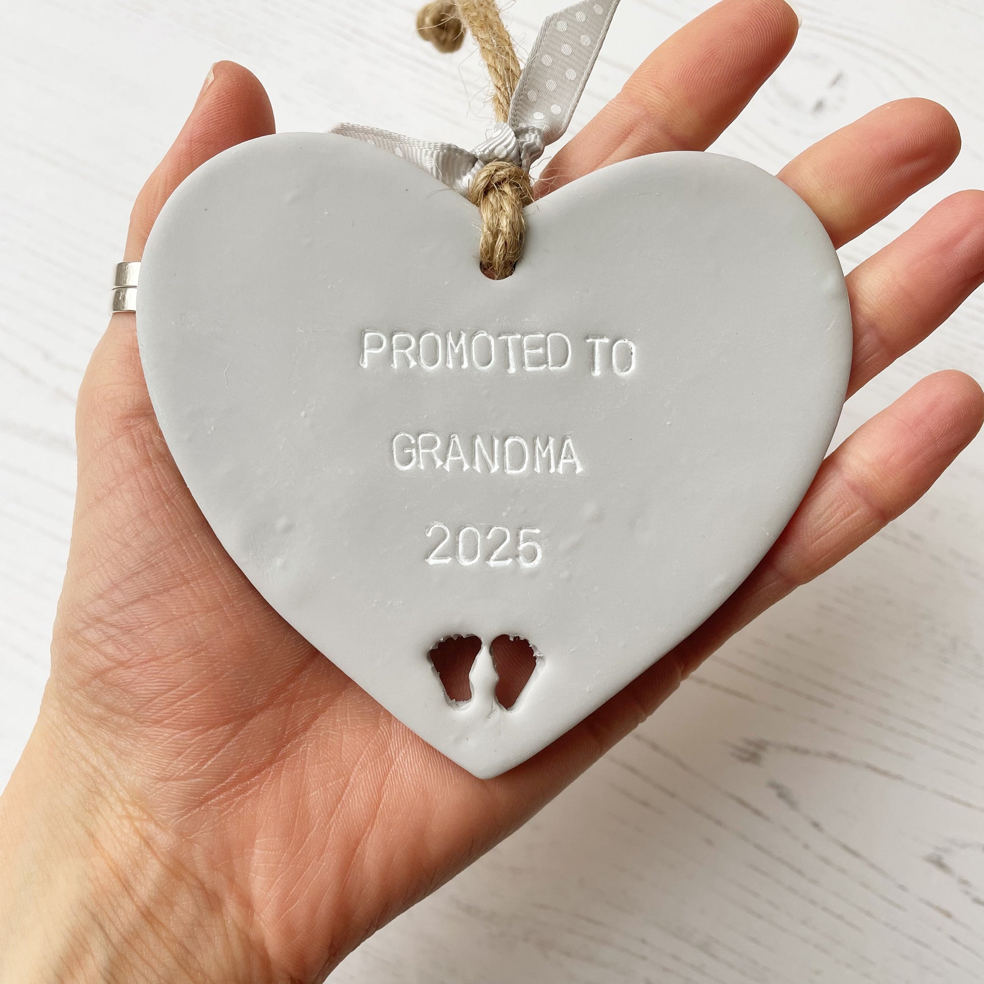 Grey clay hanging heart with baby feet cut out of the bottom and white personalisation, the heart is personalised with PROMOTED TO GRANDMA 2025