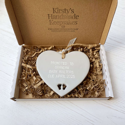 Grey clay hanging heart with baby feet cut out of the bottom and white personalisation, the heart is personalised with PROMOTED TO GRANDMA BABY WALTERS DUE APRIL 2025