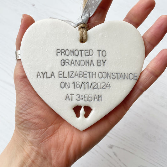 Pearlised white clay hanging heart with baby feet cut out of the bottom and grey personalisation, the heart is personalised with PROMOTED TO GRANDMA BY AYLA ELIZABETH CONSTANCE ON 16/11/2024 AT 3:55 AM