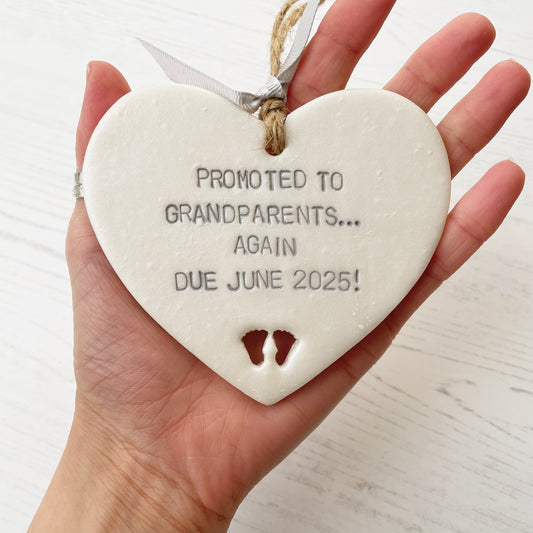 Pearlised white clay hanging heart with baby feet cut out of the bottom and grey personalisation, the heart is personalised with PROMOTED TO GRANDPARENTS… AGAIN DUE JUNE 2025!