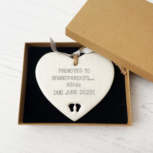 Pearlised white clay hanging heart with baby feet cut out of the bottom and grey personalisation, the heart is personalised with PROMOTED TO GRANDPARENTS… AGAIN DUE JUNE 2025!